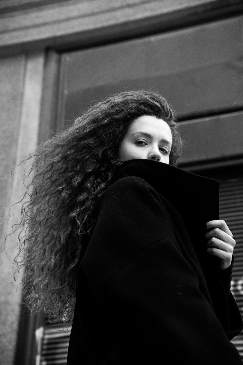 A woman with curly hair holding her coat collar up, in black and white
