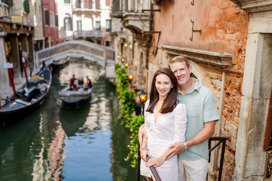 A couple standing close on a small balcony over a canal, a gondola behind them