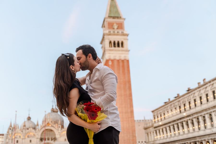 A couple kissing in St Mark's Square below the bell tower, red flowers in her hand