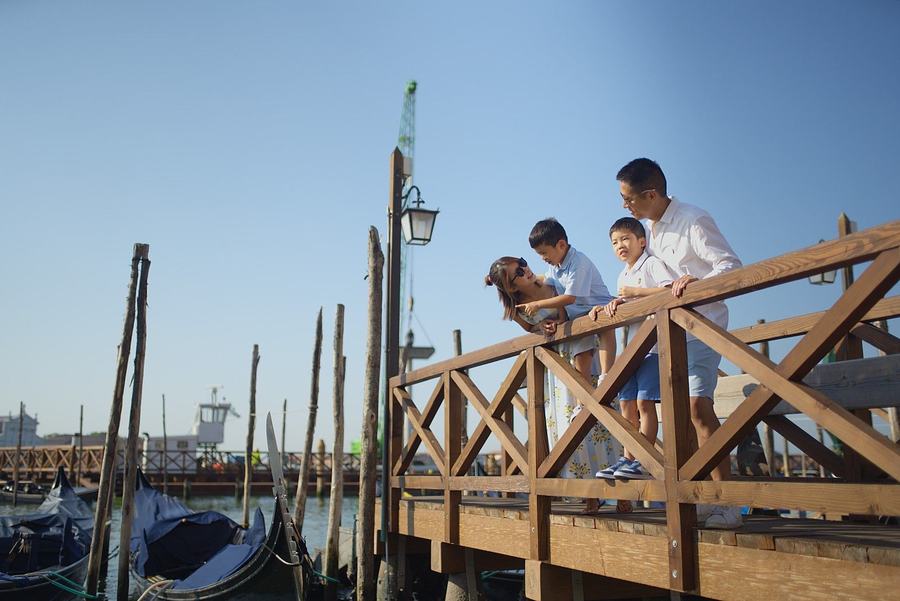 A family leaning on a wooden pier railing beside the gondolas under a blue sky