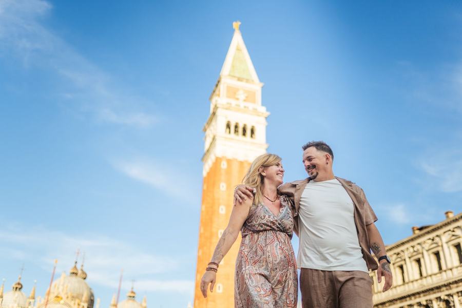 A couple walking arm in arm below the bell tower of St Mark's against a blue sky