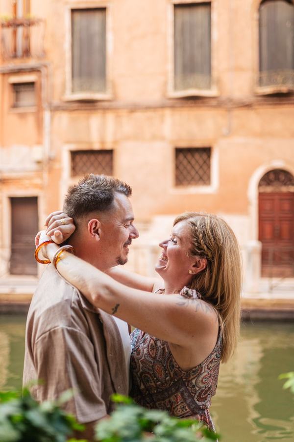 A laughing couple embracing by a canal in front of a warm orange wall