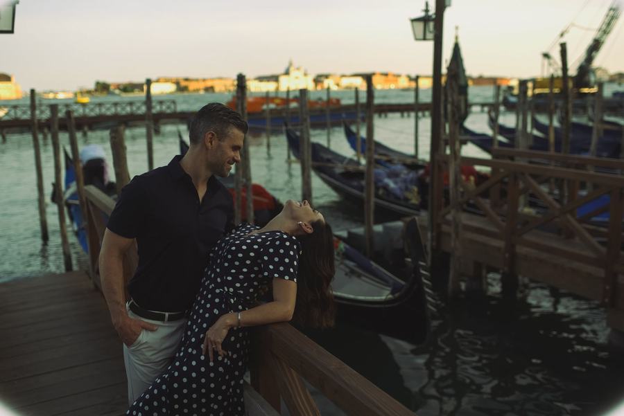 A man holding a laughing woman in a polka-dot dress by the gondolas at sunset in Venice